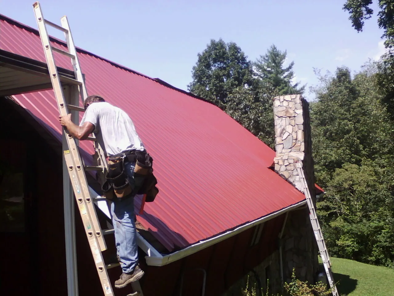 Roofer on a ladder beside a red metal roof and stone chimney — Soffit & Fascia Repair by Roofing Brothers in Zion Grove